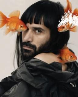 Amos Le Blanc in a dark sculptural jacket framed by three orange goldfish